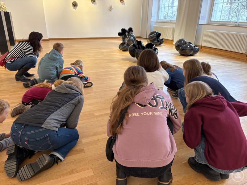 Museumspädagogik Mehrere Kinder und Erwachsene knien in einem hellen Museumsraum auf dem Holzfußboden und betrachten gemeinsam Ausstellungsstücke, darunter mehrere schwarze Figuren oder Modelle, die auf dem Boden liegen.