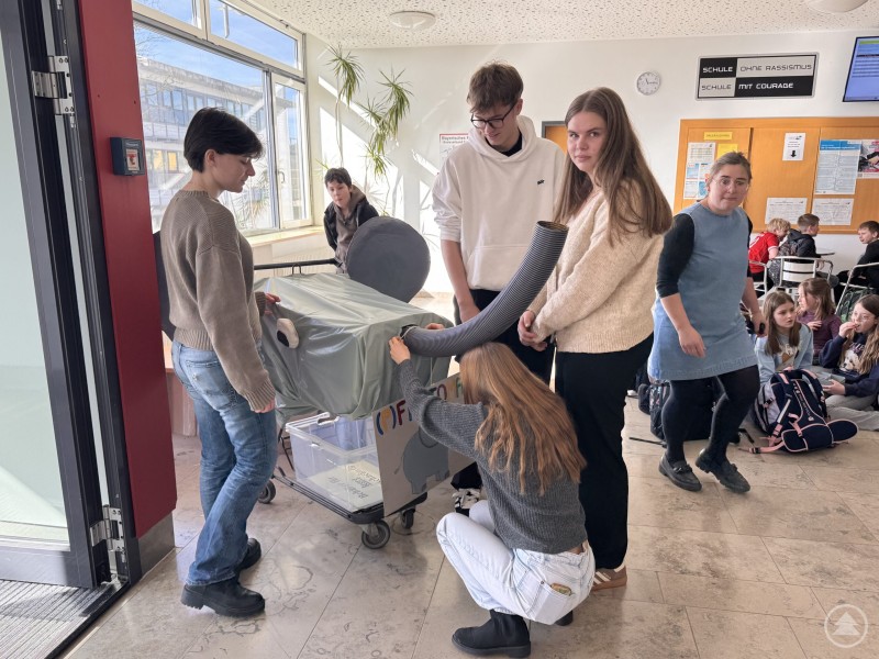 Beim Aufbau des (P)FANTOMAT wird deutlich, wie viel Teamarbeit und handwerkliches Geschick in dem Projekt steckt. Mehrere Schülerinnen und Schüler arbeiten in einem Schulflur am Aufbau des (P)FANTOMAT. Während einige die Konstruktion festhalten, montiert eine Schülerin die Verkleidung am Wagen.