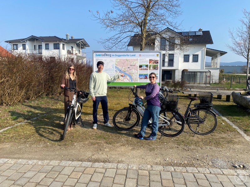 Mit vollem Einsatz für den Radtourismus: Anna-Lena Kirchner, Simon Renner und Franziska Moser von der Stadt Deggendorf präsentieren die neue Infotafel in Fischerdorf Drei Personen stehen mit Fahrrädern vor einer großen Infotafel am Isarradweg. Im Hintergrund sind Häuser und eine Landschaft zu sehen, während eine Person auf die Karte zeigt.