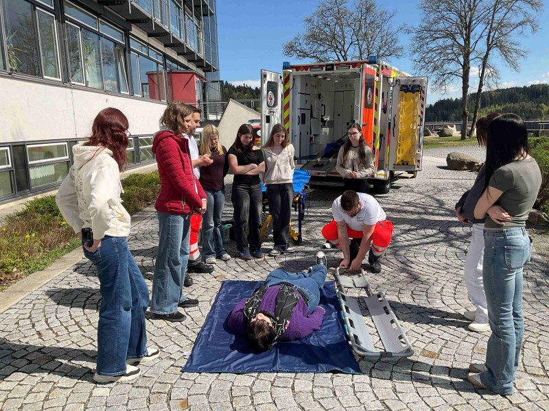 Mehrere Sch&uuml;lerinnen stehen um eine liegende Person und beobachten, wie ein Rettungssanit&auml;ter den Einsatz einer Trage erkl&auml;rt.