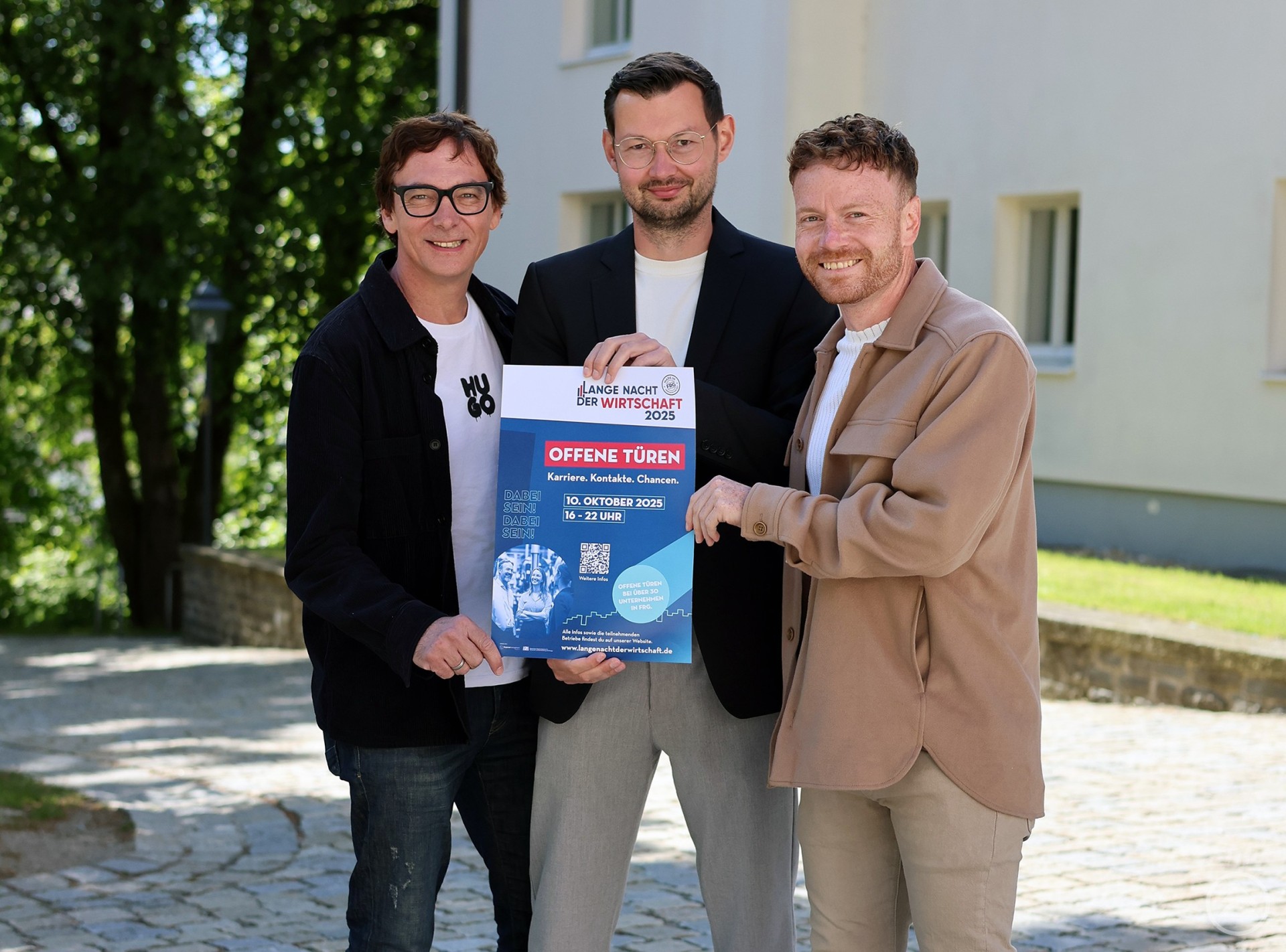 Drei Männer stehen gemeinsam im Freien und halten ein Plakat zur „Langen Nacht der Wirtschaft 2025“. Im Hintergrund ist ein Gebäude und grüne Bäume zu sehen.