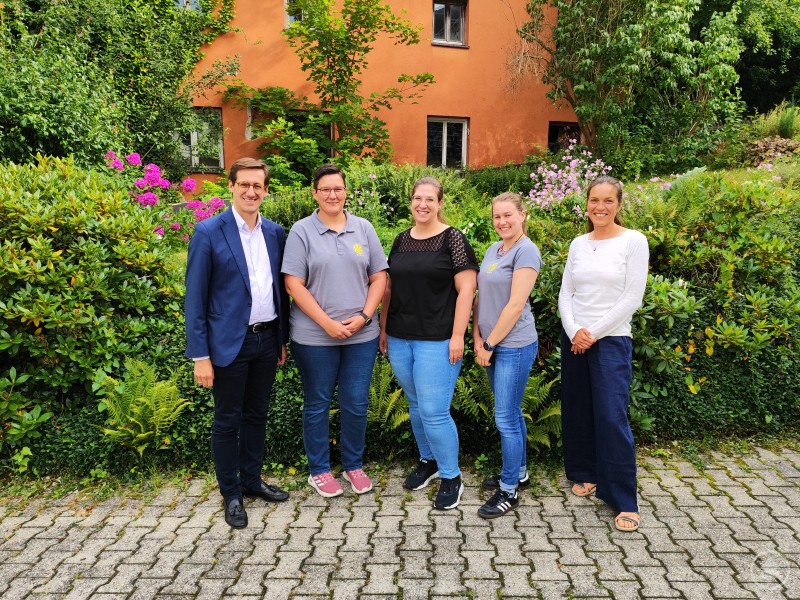 Tauschten sich über Ansätze und Notwendigkeiten einer „Zukunftsgarantie für die Jugend“ aus: (v. li. n. re.) MdL Dr. Stefan Ebner, KJR-Vorsitzende Monika Wastl, KJR-Geschäftsführerin Kathrin Zitzelsberger Fernandes, stellv. KJR-Vorsitzende Magdalena Kerscher und die pädagogische Fachkraft des KJR, Sigrid Kick. Fünf Personen – drei Frauen, ein Mann im Anzug und eine weitere Frau – stehen nebeneinander vor einer Hecke mit Blumen und einem orangefarbenen Gebäude, lächeln freundlich in die Kamera.