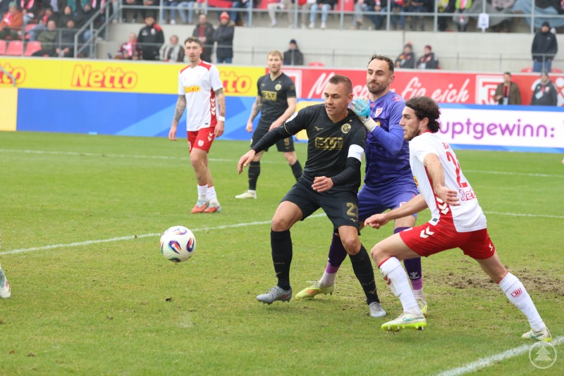 Hektische Szene im Regensburger Strafraum, Torwart Gebhardt im Einsatz gegen Rostocker Angreifer. Im Strafraum kämpfen ein Rostocker Spieler und ein Jahn-Spieler (sowie der Torwart des SSV Jahn Regensburg) um den Ball, der sich direkt vor ihnen am Boden befindet. Weitere Spieler beobachten die Szene.