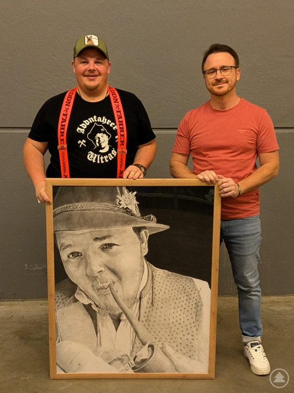 Florian Eibl überreichte dem Addnfahrer sein handgezeichnetes Portrait persönlich. Der Portraitzeichner Florian Eibl und der bayerische Comedian Addnfahrer stehen nebeneinander und präsentieren ein großes gerahmtes Schwarzweiß-Portrait.