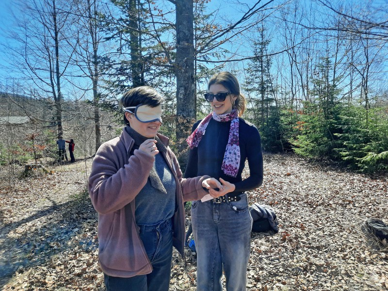 Beim Einführungsseminar standen unter anderem Methoden der Umweltbildung im Fokus. Eine Teilnehmerin mit Schlafmaske probiert bei einem Workshop im Wald eine Übung der Umweltbildung aus, daneben steht eine weitere junge Frau und begleitet die Situation.
