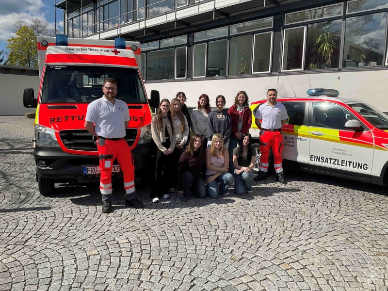 Eine Gruppe von Sch&uuml;lerinnen steht gemeinsam mit zwei Rettungskr&auml;ften vor einem Rettungswagen und einem Einsatzfahrzeug.