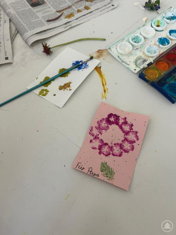 Auf einem Tisch liegen Wasserfarben, Pinsel, Bl&auml;tter und selbst gestaltete Karten mit Bl&uuml;ten- und Farbmotiven.