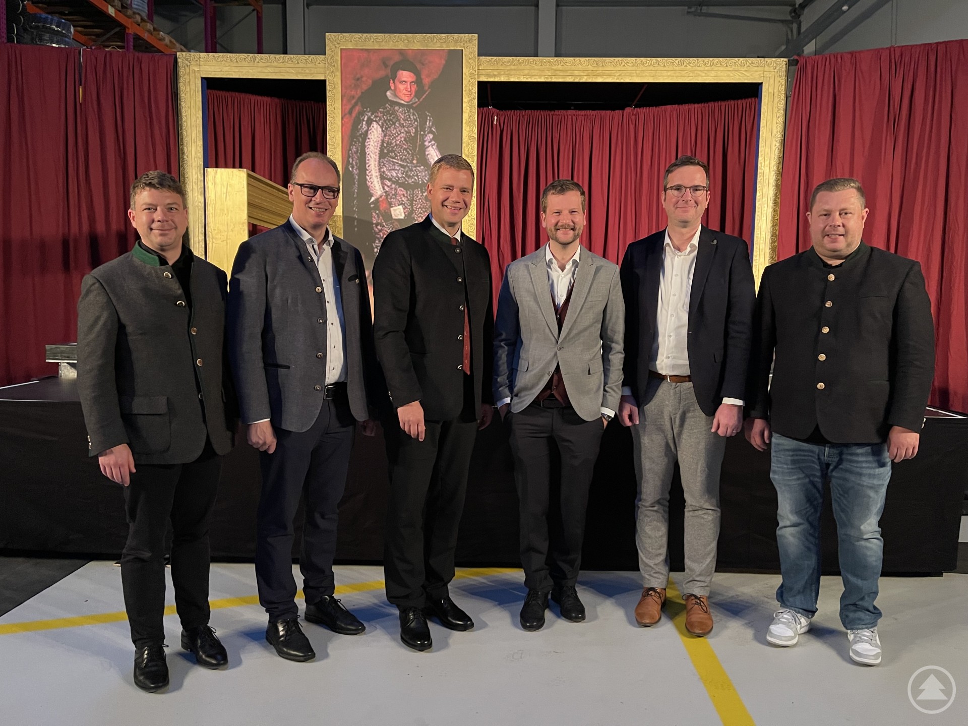 Freuten sich über die gelungene Premiere des Kulturmobils in Bodenmais: Pfarrer Alexander Kohl (von links), Abgeordneter Martin Behringer, MdL, Bezirkstagspräsident Dr. Olaf Heinrich, Intendant und Kulturreferent Dr. Laurenz Schulz, Bezirksheimatpfleger Dr. Clemens Knobling und der Bürgermeister von Bodenmais, Michael Adam. Sechs Männer in Anzügen und Trachten stehen vor einem bühnenartigen Hintergrund mit roten Vorhängen und einem großen gerahmten Gemälde.