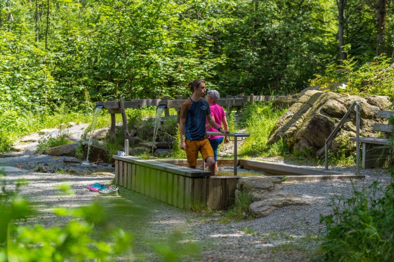 Ein junger Mann und eine &auml;ltere Frau nutzen ein weiteres Kneippbecken inmitten eines naturbelassenen, sonnigen Waldareals mit Holzrinnen und Wasserzul&auml;ufen.