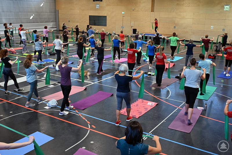 Unter dem Motto „Einfach Frau – Einfach stark“ kamen zahlreiche Frauen in Freyung zusammen, um etwas für ihre Gesundheit zu tun. Viele Frauen unterschiedlichen Alters trainieren gemeinsam in einer Sporthalle. Sie stehen auf Gymnastikmatten und arbeiten mit grünen Fitnessbändern, während eine Übungsleiterin vorne auf einer Bühne anleitet.