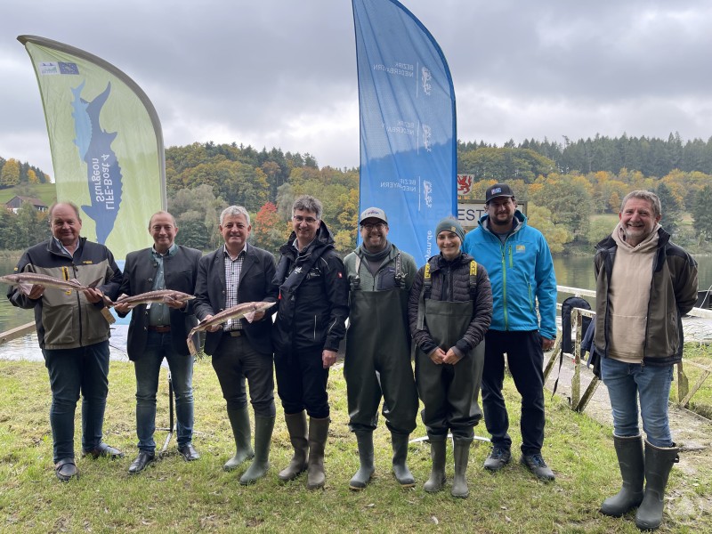 Nahmen am Besatztermin teil: Michael Kreiner, Präsident Fischereiverband Niederbayern (v. l.), Franz Langer, Bürgermeister des Marktes Windorf, Ministerialrat Dr. Reinhard Reiter, Dr. Stephan Paintner, Leiter der Fischereifachberatung des Bezirks Niederbayern, Projektleiter LIFE-Boat for Sturgeons Dr. Thomas Friedrich, Mitarbeiterin LIFE-Boat f.S. Heidrun Eichhorn, Dominik Bernolle, Landesamt f. Umwelt und Ralf Eibl, Vorstand Bezirksfischereiverein Passau. Acht Personen stehen am Donauufer vor Bannern des Bezirks Niederbayern und des Projekts LIFE-Boat for Sturgeons. Einige halten Sterlets in den Händen.