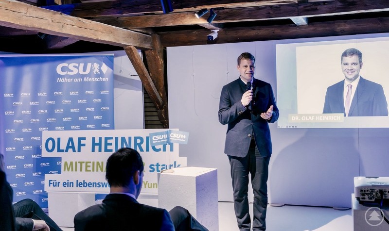 Dr. Olaf Heinrich bei seiner Ansprache zur Bürgermeister-Nominierung im Europahaus. Ein Mann im Anzug spricht auf einer Bühne mit Mikrofon. Hinter ihm ist eine Leinwand mit seinem Porträt und ein Banner der CSU. Im Vordergrund sind Zuhörer zu sehen, links steht ein großes Schild mit seinem Namen.