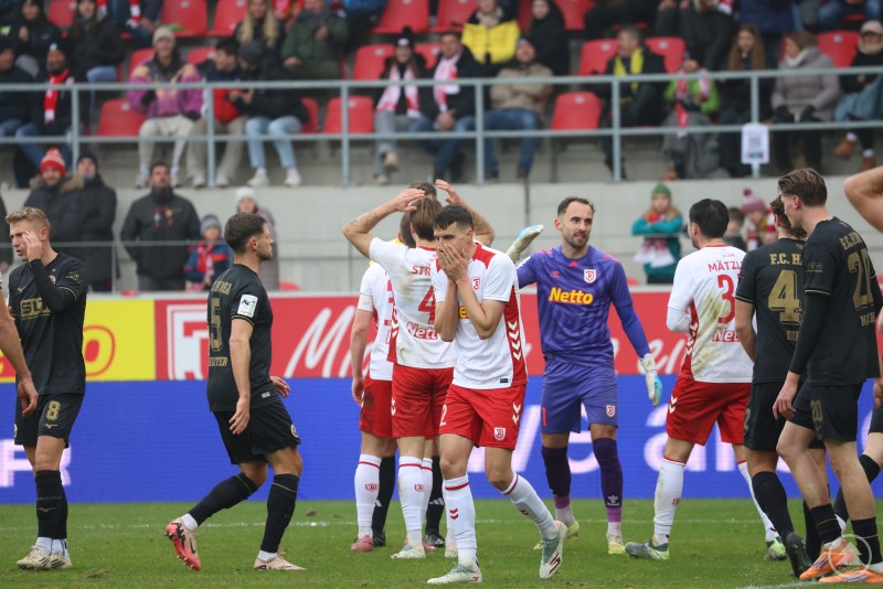 Mehrere Spieler des SSV Jahn Regensburg und von Hansa Rostock stehen auf dem Feld, teils sichtlich enttäuscht oder nachdenklich, einer hält sich das Gesicht vor die Hände. Im Hintergrund sind weitere Spieler und Zuschauer zu sehen.