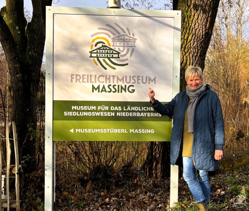 Betriebsleiterin Silke Holzner hat mit ihrem Team alles vorbereitet für den Start in das neue Museumsjahr Betriebsleiterin Silke Holzner steht vor einem großen Schild des Freilichtmuseums Massing und zeigt lächelnd auf das Museumslogo.