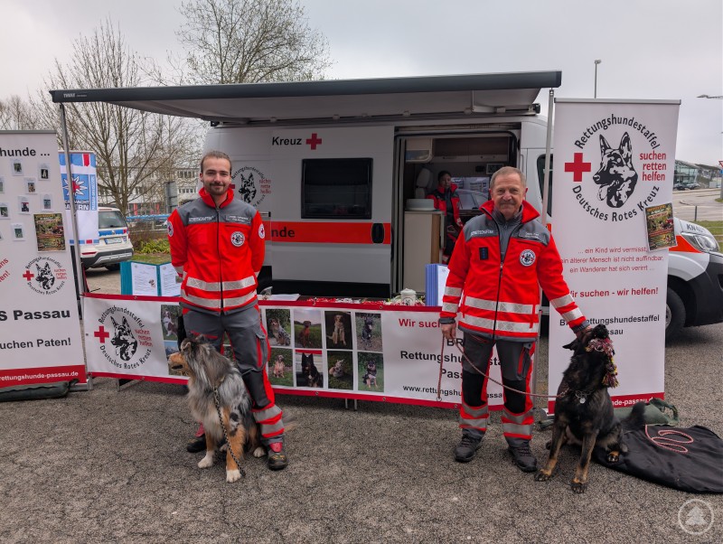 Zwei der ehrenamtlichen Rettungshundestaffelf&uuml;hrer posieren zusammen mit ihren Hunden vor dem Messestand.