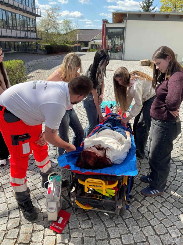 Mehrere Sch&uuml;lerinnen stabilisieren gemeinsam eine Person auf einer Trage, w&auml;hrend ein Rettungssanit&auml;ter die Handgriffe erkl&auml;rt.