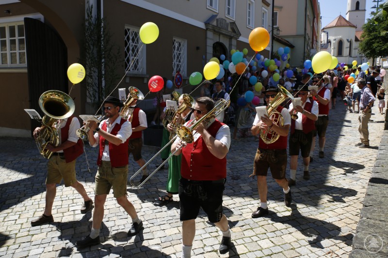 Gleich trägt sich die Tuba am Kinderzug etwas leichter mit Ballon. Eine Blaskapelle in Tracht marschiert durch eine gepflasterte Straße, begleitet von bunten Luftballons und einem großen Publikum.