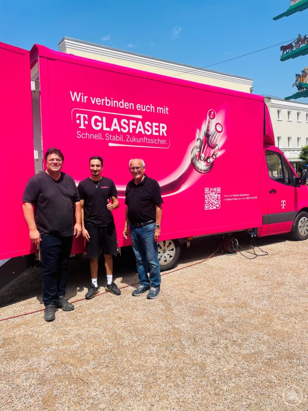 v. l. MGR Dr. Andreas Geiß, Herr Aleko, Glasfaserbotschafter der Deutschen Telekom, und 2. Bürgermeister Günter Klampfl vor dem Info-Mobil in Schönberg. Drei Männer stehen vor einem auffällig pinken Info-Mobil der Deutschen Telekom mit der Aufschrift „Wir verbinden euch mit Glasfaser“. Im Hintergrund ist ein Maibaum zu erkennen, der Ort wirkt sommerlich.