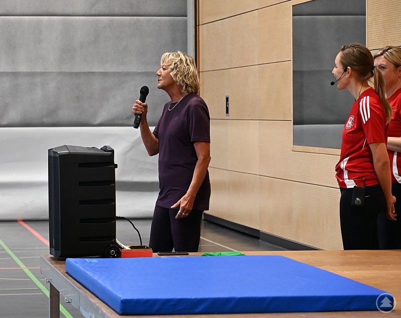 Stellvertretende Landrätin Hilde Greiner bei der Begrüßung der Teilnehmerinnen. Eine Frau mit Mikrofon spricht zu den Teilnehmerinnen, daneben stehen zwei junge Frauen in roten Sportshirts und hören aufmerksam zu.