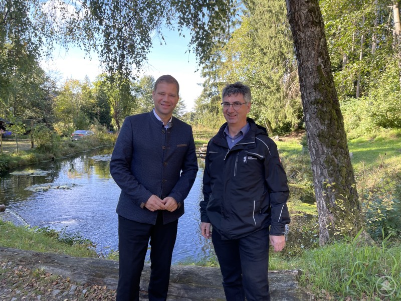 Bezirkstagspräsident Dr. Olaf Heinrich und Dr. Stephan Paintner, Leiter der Fischereifachberatung des Bezirks, besuchten zum „Tag der offenen Fischzucht“ das Fischzucht Unikum in Hunding. Zwei Männer stehen am Rand eines idyllischen Fischteichs, im Hintergrund Bäume und Wasserpflanzen. Beide lächeln in die Kamera, einer trägt eine Trachtenjacke, der andere eine schwarze Outdoor-Jacke.