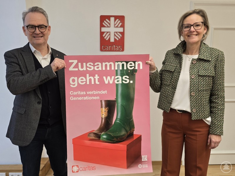 Die Passauer Caritasvorstände Andrea Anderlik und Stefan Seiderer freuen sich auf den Kampagnenstart in Passau. Das Plakatmotiv stammt ebenfalls aus Passau. Die Stiefel der Kita-Leiterin Constanze Simmel und eines Kindes symbolisieren das Miteinander von Jung und Alt. Zwei Personen, ein Mann und eine Frau, stehen in einem hellen Raum vor dem Caritas-Logo und halten gemeinsam ein großes pinkes Plakat mit der Aufschrift „Zusammen geht was. Caritas verbindet Generationen“. Auf dem Plakat sind zwei Paar Gummistiefel – ein großes grünes und ein kleines mit Tiermotiv – zu sehen. Beide lächeln in die Kamera.