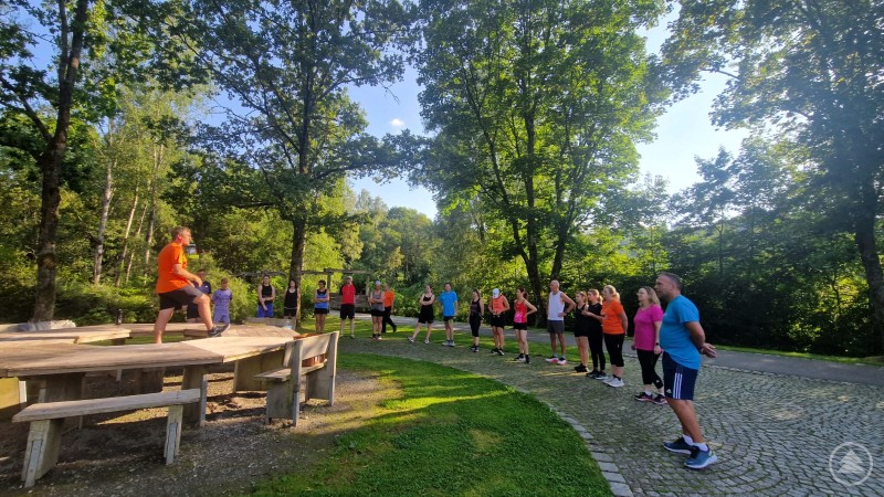 Das Bild zeigt eine sportliche Gruppe von etwa 20 Personen, die sich bei sonnigem Wetter im Grünen zu einem Outdoor-Training versammelt hat. Im Mittelpunkt steht ein Trainer in orangefarbenem Shirt, der auf einem Holzpodest eine Übung demonstriert. Die Teilnehmenden – in Sportkleidung – hören aufmerksam zu und stehen auf einem gepflasterten Weg in einem parkähnlichen Waldstück. Es handelt sich wahrscheinlich um ein gemeinsames Lauf- oder Fitnessprogramm, möglicherweise im Rahmen einer Aktion wie LAUF10!, bei der Einsteiger behutsam an Ausdauersport herangeführt werden. Die Atmosphäre ist motivierend und gemeinschaftlich.