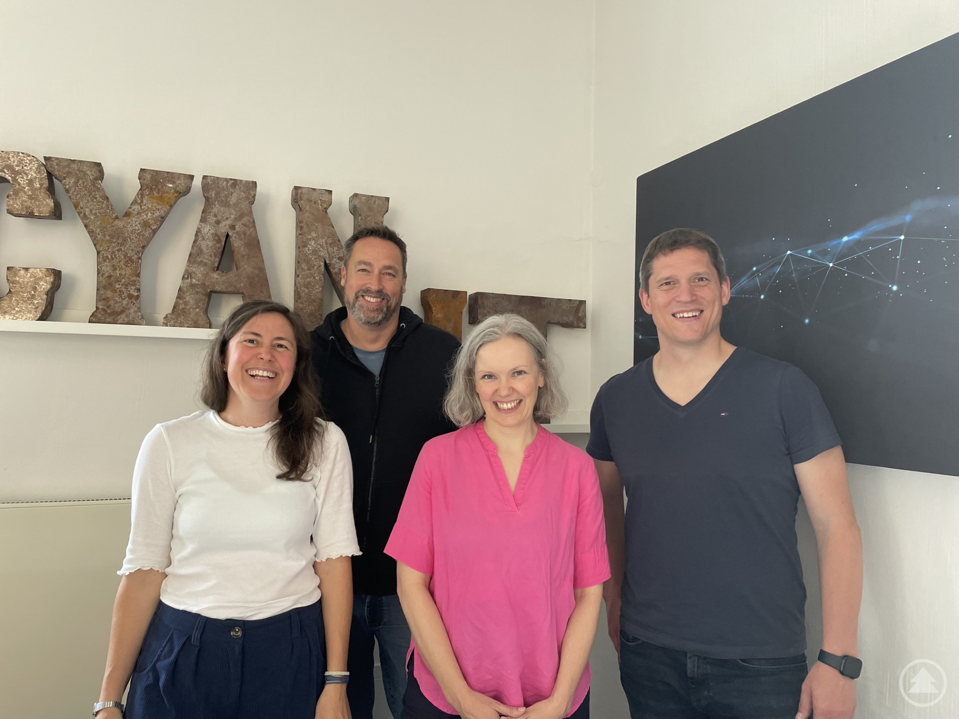 Vier lächelnde Personen, zwei Männer und zwei Frauen, stehen in einem modernen Büro vor einer Wand mit dem Schriftzug 'CYAN' und einer abstrakten Grafik.