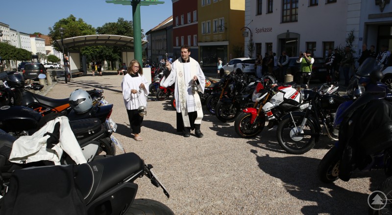Motorräder wurden mit Weihrauchsegen im Risiko abgeregelt. Ein Priester und eine Ministrantin segnen auf einem Platz vor der Sparkasse eine Vielzahl geparkter Motorräder.