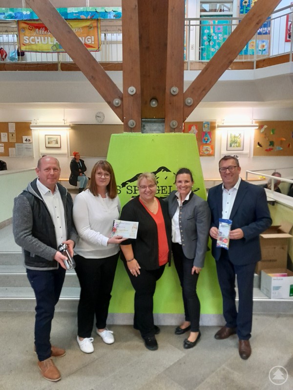 Vier Erwachsene und eine Schulleiterin stehen in der Aula der Grundschule Spiegelau vor einer grünen Wand mit Schullogo. Zwei Personen halten Geschenke für die ABC-Schützen in den Händen.