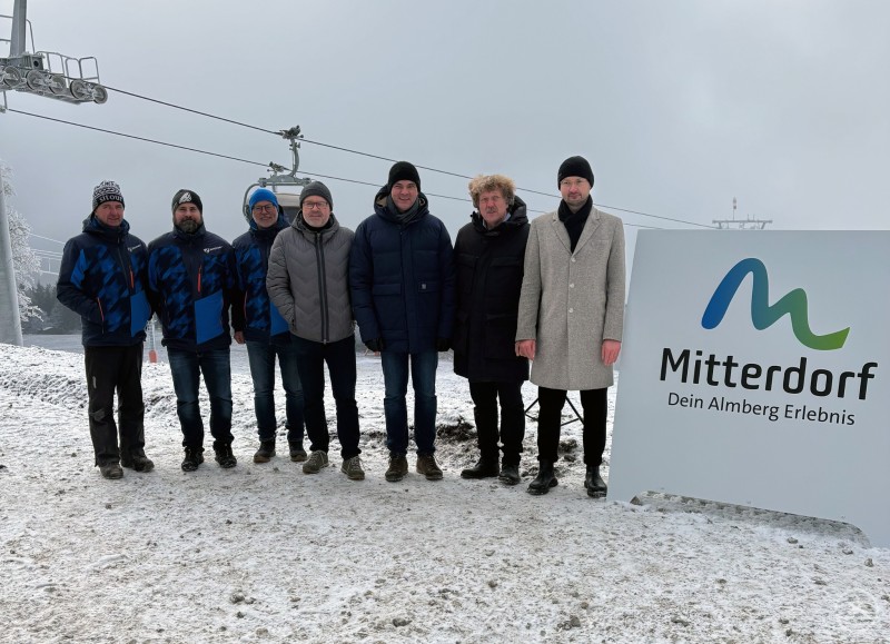 Bei der Präsentation des neuen Logos auf der Bergstation: Thomas Schrottenbaum (Betriebsleiter Mitterdorf-Liftanlagen), Marco Denk (stv. Geschäftsführer Zweckverband Wintersportzentrum Mitterdorf), Bernhard Hain (Geschäftsführer Zweckverband Mitterdorf), Grafikdesigner Willi Weber, Landrat Sebastian Gruber, Philippsreuts Bürgermeister Helmut Knaus und Rainer Jungwirth von der Agentur siimple. Eine Gruppe von sieben Männern steht im winterlichen Außenbereich vor einer Seilbahnstation. Alle tragen Winterkleidung. Rechts im Bild befindet sich ein großes Schild mit dem neuen Logo und Claim „Mitterdorf – Dein Almberg Erlebnis“. Die Umgebung ist verschneit und nebelig.
