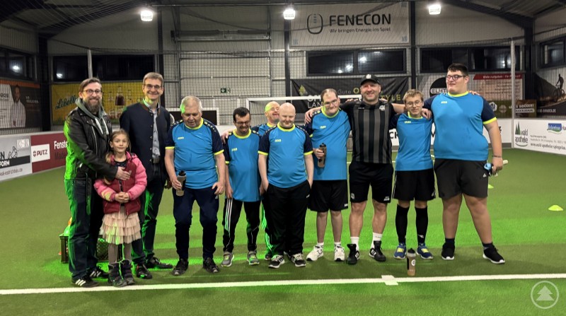 Die Bolzrebellen beim ersten Training im Bolzwerk Spiegelau zusammen mit den Initiatoren und Spiegelaus Bürgermeister: (von rechts) Fabian Kroiß, Lukas Schwarz, Michael Miedl (Eigentümer Bolzwerk), Markus Blach, Fernando Harz, Helmut Ranzinger, Patrick Poxleitner, August Czernoch. Bürgermeister Karlheinz Roth und Christian Fiebig (Landratsamt). Eine gemischte Gruppe aus Spielern und Verantwortlichen steht in einer Fußballhalle auf Kunstrasen. Die Mannschaft trägt einheitliche blaue Trikots. Neben den Spielern sind Initiatoren und Vertreter aus Politik und Organisationen zu sehen. Alle blicken lächelnd in die Kamera, die Stimmung wirkt gemeinschaftlich und positiv.
