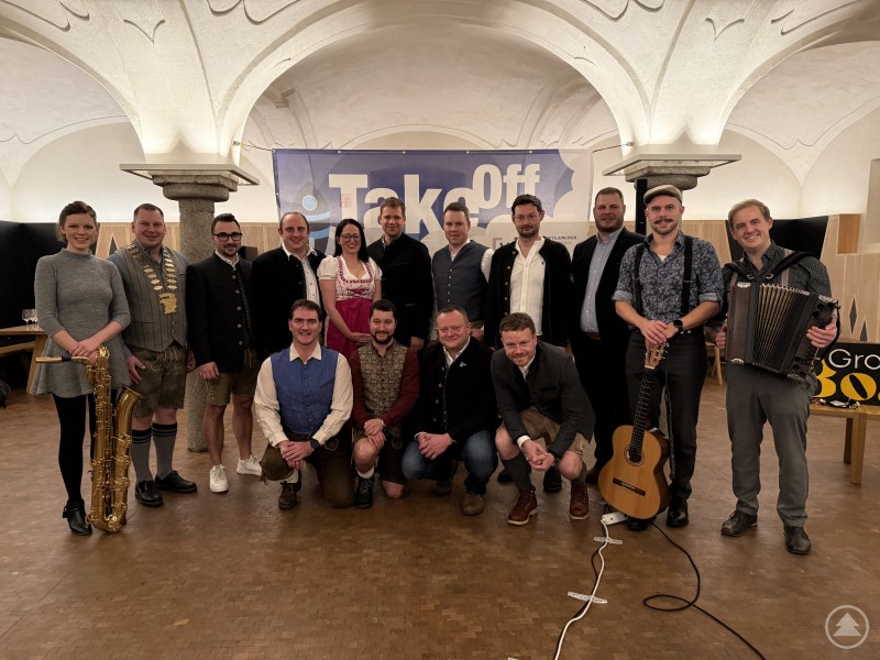 Das Organisationsteam und musikalische Begleitung beim TakeOff 2026 der Wirtschaftsjunioren Bayern in der Volksmusikakademie Freyung. Eine größere Gruppe von Menschen, überwiegend in Tracht oder festlicher Kleidung, posiert gemeinsam für ein Gruppenfoto in einem historischen Gewölbesaal. Im Hintergrund ist ein großes Banner mit der Aufschrift „TakeOff“ zu sehen. Einige Personen halten Musikinstrumente wie Saxophon, Gitarre und Akkordeon, was auf eine musikalische Begleitung hinweist. Die Stimmung wirkt ausgelassen und feierlich.