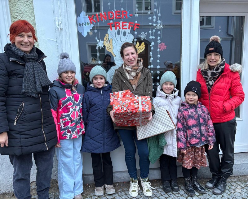 Sieben Personen, darunter vier Kinder und drei Erwachsene, stehen lachend vor einem Fenster mit der Aufschrift &bdquo;Kinder-Treff&ldquo;. Zwei Kinder und eine Frau halten bunt verpackte Geschenke in den H&auml;nden. Alle sind winterlich gekleidet.