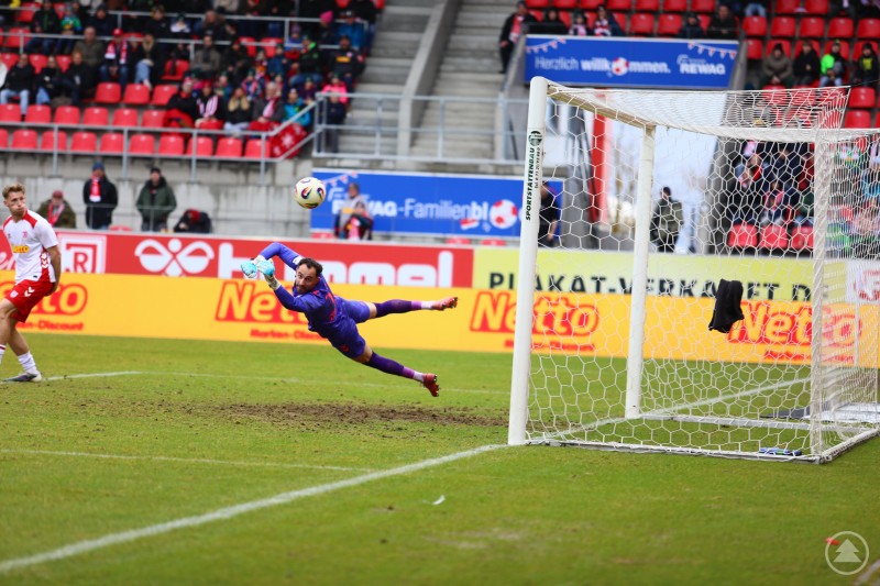Der Torwart des SSV Jahn Regensburg fliegt gestreckt durch den Strafraum und versucht einen Ball neben dem rechten Pfosten abzuwehren, im Hintergrund sind rote Trib&uuml;nen zu sehen.