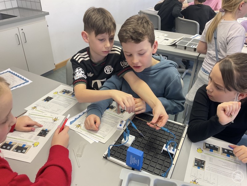 Viertklässler experimentieren beim Forschernachmittag am LLG mit elektrischen Schaltungen. Mehrere Grundschulkinder sitzen gemeinsam an einem Tisch und bauen mit Kabeln und Bauteilen einen einfachen Stromkreis auf. Arbeitsblätter und Experimentiermaterial liegen auf dem Tisch.