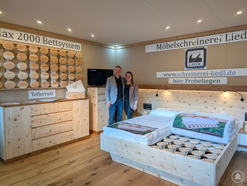 Der Geruch von Zirbenholz lag in der Luft bei Schreinerei Liedl Ein aufwendiger Messestand aus hellem Zirbenholz stellt ein komplettes Schlafzimmer dar. Die beiden Aussteller der Schreinerei Liedl stehen lächelnd neben dem Bett.