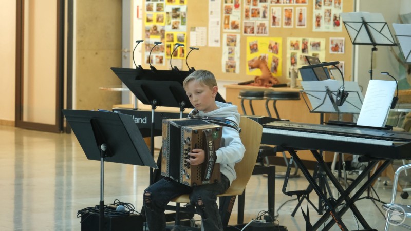 Ein junger Sch&uuml;ler sitzt mit einer Harmonika auf einer B&uuml;hne und spielt konzentriert vor Notenst&auml;ndern und Instrumenten.