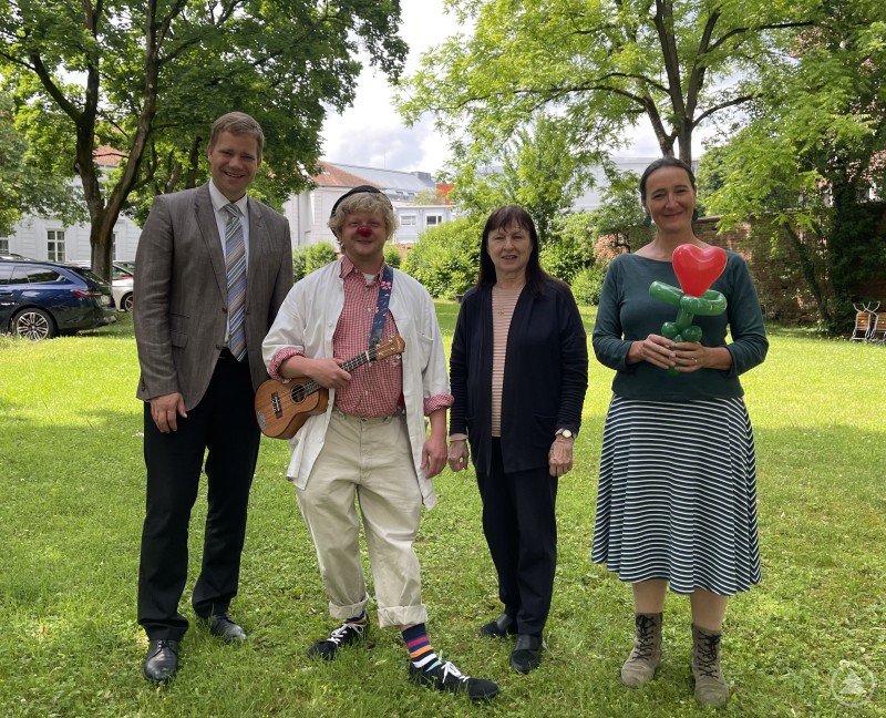 Bezirkstagspräsident Dr. Olaf Heinrich (links) und Kulturreferentin Veronika Keglmaier (rechts) freuten sich über den Besuch von KlinikClown Muck und der Vereinsvorsitzenden Elisabeth Makepeace-Vondrak. Vier Personen stehen auf einer Wiese im Grünen, darunter ein Klinikclown mit roter Nase und Ukulele, eine Frau mit Luftballonfigur, ein Mann im Anzug und eine weitere Frau in dunkler Kleidung.