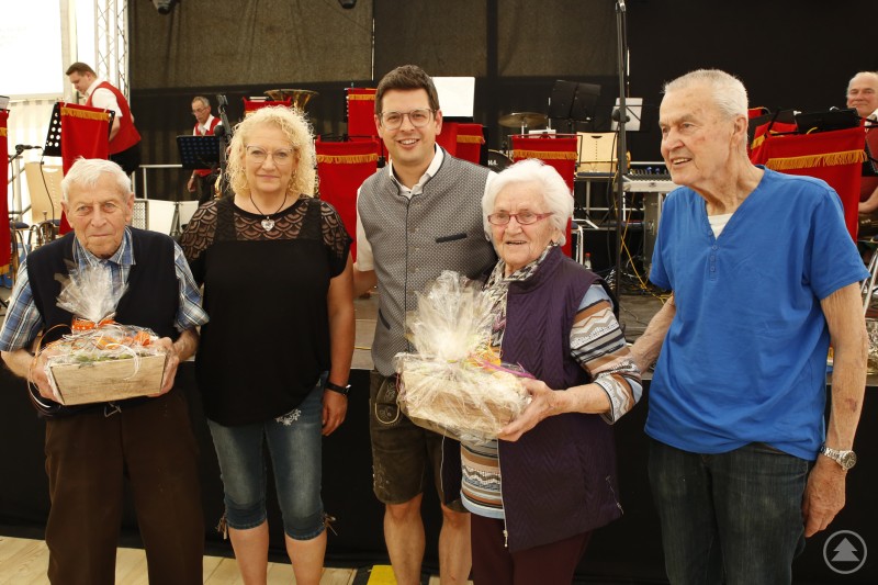 Aus den 30ern stammen Mr. und Mrs. Volksfest, Maria Buchecker und Bepperl Murr, denen Seniorenbeauftrage Doris Rötzer und Bürgermeister Martin Pichler Geschenkkörbe überreichten. Zwei Senioren und eine Seniorin halten Geschenkkörbe, neben ihnen stehen eine Frau und ein Mann in Tracht. Im Hintergrund eine Bühne mit Musikanten.