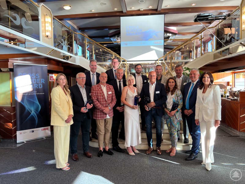 Gruppenfoto der Preisträgerinnen und Preisträger sowie Organisatoren des Cross Border Awards 2025 an Bord des Galaschiffes „Regina Danubia“ der Donauschifffahrt Wurm & Noé. Eine Gruppe von etwa 17 festlich gekleideten Personen steht auf einem Schiff in einem eleganten Innenbereich für ein Gruppenfoto zusammen. Im Hintergrund ist ein Bildschirm mit dem Schriftzug „CROSS BORDER AWARD“ sowie ein Banner mit gleichem Titel zu sehen. Einige Personen halten Glas-Trophäen in der Hand.