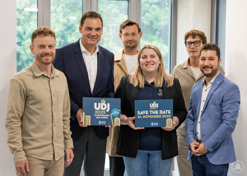 Das Organisationsteam des Preises „Unternehmen des Jahres“ 2025 freut sich auf zahlreiche Bewerbungen aus der Region: Leiter Stabsstelle Kreisentwicklung Johannes Gastinger, Landrat Sebastian Gruber, Wirtschaftsjunioren Past President Rainer Jungwirth, Wirtschaftsjunioren Vizepräsidentin Anna Schraml, Regionalmanager Stefan Schuster, Wirtschaftsjunioren Präsident Alexander Treml. Sechs Personen stehen in einer hellen Eingangshalle und blicken lächelnd in die Kamera. Zwei von ihnen halten Schilder mit dem Logo „UDJ“ und dem Hinweis „Save the Date“.