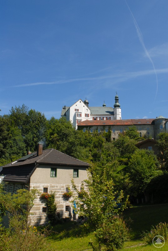 Historisches Schloss mit weißen Fassaden und Türmen erhebt sich über einer bewaldeten Anhöhe.