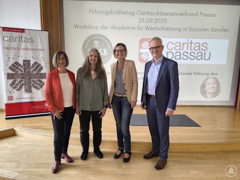 Vier Personen stehen nebeneinander auf einer B&uuml;hne, im Hintergrund ist ein Pr&auml;sentationsscreen mit Caritas-Logos zu sehen.