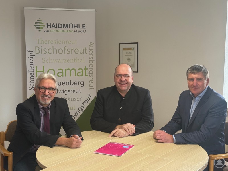 Bürgermeister Roland Schraml, Markus Münch von der Deutschen Telekom und ein weiterer Vertreter im Gespräch zum Glasfaserausbau in Haidmühle. Drei Männer sitzen an einem runden Holztisch in einem Besprechungsraum. Hinter ihnen steht ein Roll-up mit dem Logo der Gemeinde Haidmühle und verschiedenen Ortsnamen. Auf dem Tisch liegt eine pinkfarbene Informationsmappe. Alle drei tragen Businesskleidung und blicken in die Kamera.