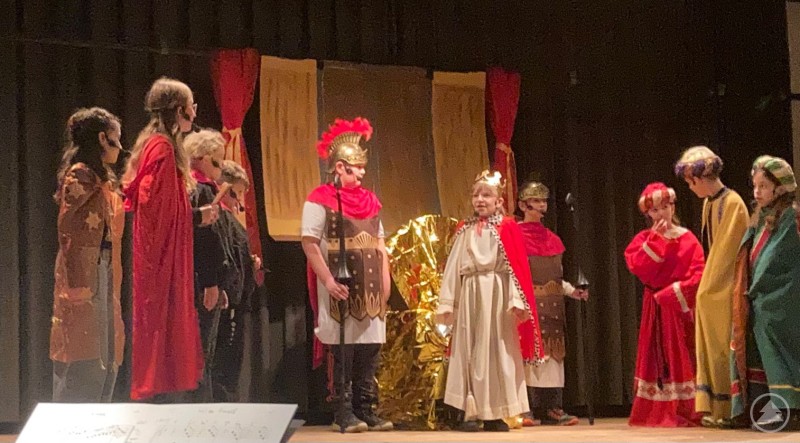 König Herodes lauscht den Worten der Heiligen Drei Könige und seiner Sterndeuter. Kinder der Grundschule Spiegelau stehen auf einer Bühne in historischen Kostümen. Zu sehen sind König Herodes, die Heiligen Drei Könige, Palastwachen und weitere Figuren einer Musicalaufführung mit weihnachtlichem Hintergrund.