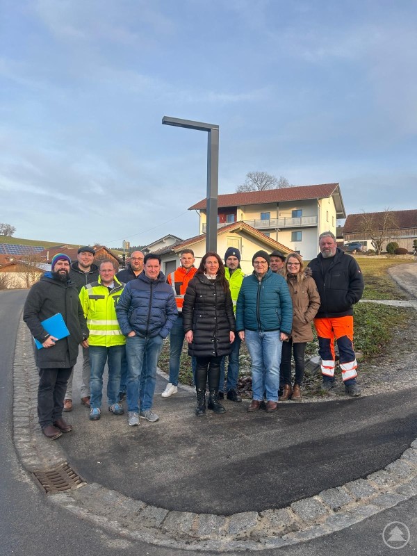 Schlussabnahme für den ersten Bauabschnitt im Rahmen der Dorferneuerung Gehmannsberg: Vertreter von Gemeinde, Verbänden und Baufirma am neuen Straßenabschnitt. Eine Gruppe von zwölf Personen steht im Freien auf einer neu asphaltierten Straße vor einem modernen Straßenbeleuchtungsmast. Im Hintergrund sind Wohnhäuser und ein leicht ansteigender Hang zu sehen. Die Menschen tragen wetterfeste Kleidung, einige sind in Warnwesten oder Arbeitskleidung, andere in Freizeit- oder Bürokleidung. Die Stimmung ist freundlich und sachlich.