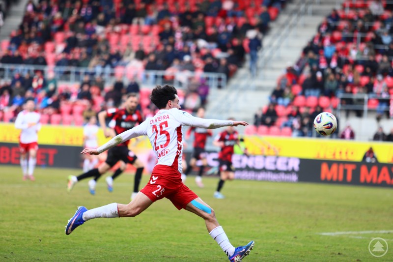 Ein Regensburger Spieler mit der R&uuml;ckennummer 25 holt zum Schuss aus, w&auml;hrend Gegenspieler in schwarz-rot heranst&uuml;rmen.