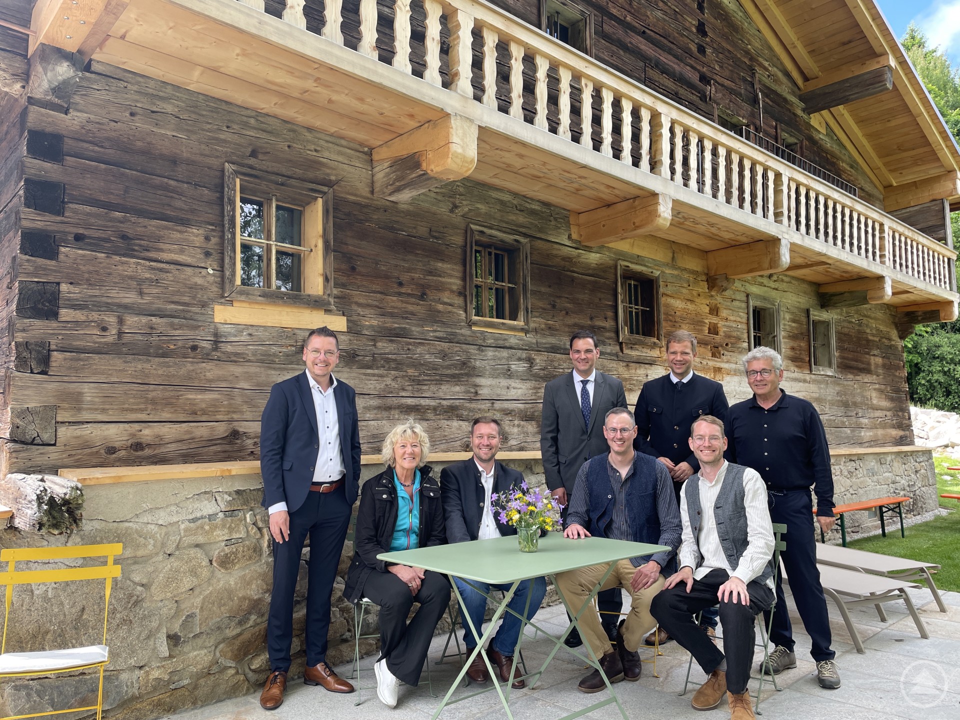 Florian Hartl und Florian Gugenhan (sitzend v.r.) haben den Staindlhof saniert. Bei der Eröffnung konnten sich (v.l.) Waldkirchens Bürgermeister Heinz Pollack, stv. Landrätin Hilde Greiner, MdL Josef Heisl, Landrat Sebastian Gruber, Bezirkstagspräsident Dr. Olaf Heinrich und Architekt Norbert Paukner von der neuen Qualität des Baudenkmals überzeugen. Eine Gruppe von acht Personen, darunter Frauen und Männer, steht oder sitzt vor einem großen, traditionellen Holzhaus mit Fachwerk und kleinen Fenstern, das von einer Sanierung zeugt.