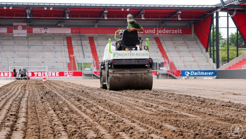 Eine Walze der Firma Richter Rasen bearbeitet den Boden eines Fußballstadions, im Hintergrund Tribünen mit roten Akzenten.