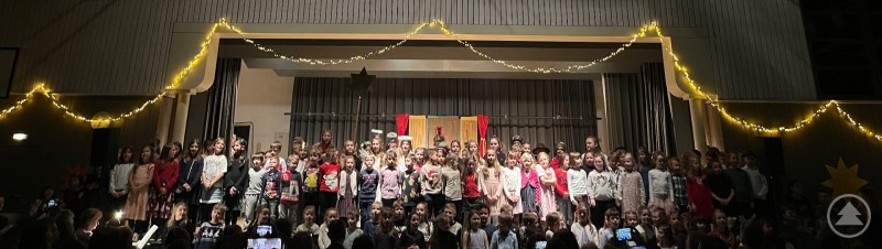Alle Kinder singen gemeinsam das Lied „Und wenn er wirklich wiederkäm“. Alle Schülerinnen und Schüler der Grundschule Spiegelau stehen gemeinsam auf der festlich geschmückten Bühne der Turnhalle und singen ein Abschlusslied, während im Publikum zahlreiche Zuschauer das Weihnachtsmusical verfolgen.